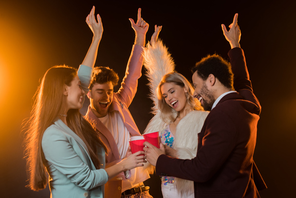 group of excited multicultural friends toasting plastic cups and rejoicing during party on black - Photo, Image