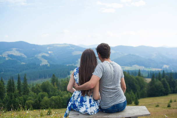 Back view of Romantic young couple enjoy the view of the mountains sitting on a bench on a sunny day in Carpatians. Loving couples date. Romantic outdoors. - Фото, зображення