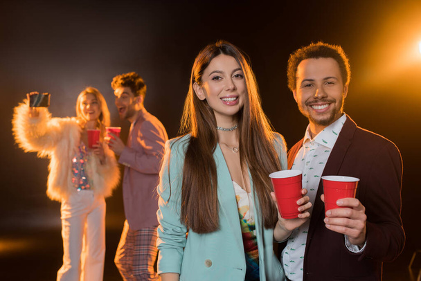 cheerful brunette woman and african american man holding plastic cups near friends taking selfie during party on black - Foto, afbeelding