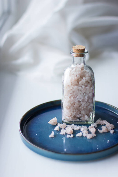 Pink sea salt in glass bottle on the blue plate. SPA body care concept. Bath salts, glass bottles with pink sea salt on white background. - Photo, Image