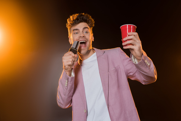man singing while holding microphone and plastic cup on black - Fotoğraf, Görsel