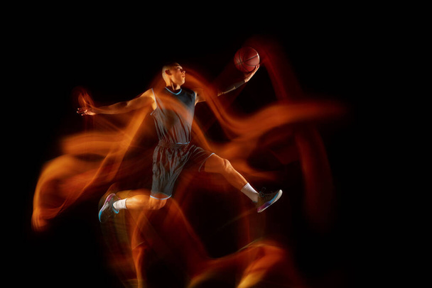 Young east asian basketball player in action and jump in mixed light over dark studio background. Concept of sport, movement, energy and dynamic, healthy lifestyle. - Zdjęcie, obraz