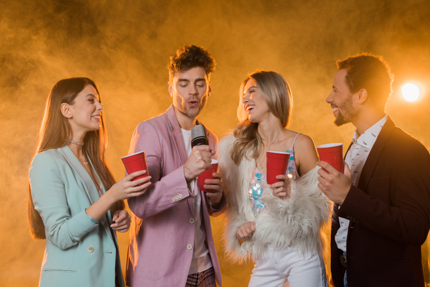 man singing while in microphone near multicultural friends with plastic cups on black with smoke - Foto, Imagem