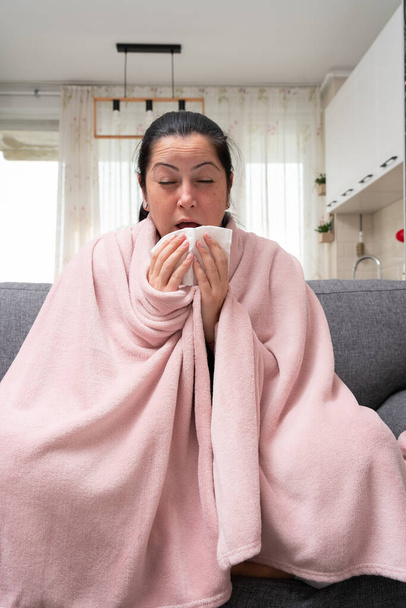 Kobieta z zakażeniem zatok kichająca trzymająca serwetkę w dłoni jako objaw zakażenia wirusem grypy lub przeziębienia siedzącą na kanapie - Zdjęcie, obraz