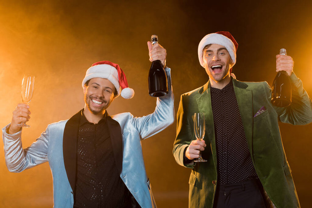 excited interracial men in santa hats holding bottles of champagne and glasses on black  - Fotografie, Obrázek