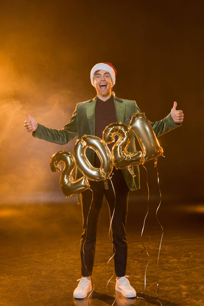 サンタ・ハットの興奮した男が2021年の数字で風船の近くに親指を立て  - 写真・画像