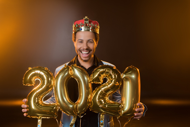 happy african american man in crown holding balloons with 2021 numbers on black  - Foto, afbeelding