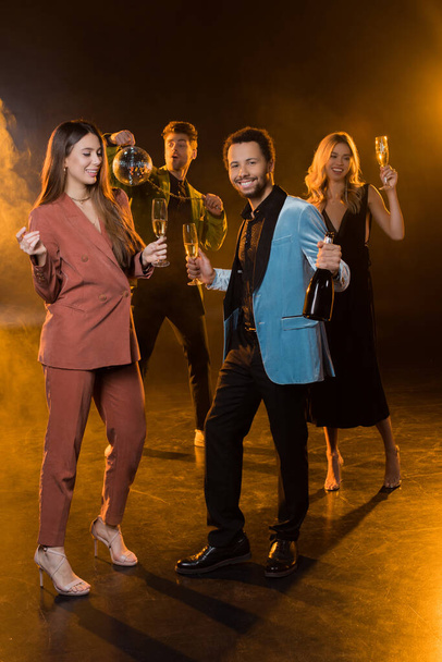 full length of happy multicultural couple holding glasses of champagne and bottle near friends on blurred and black background - Photo, Image