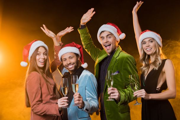 cheerful multicultural friends in santa hats holding glasses with champagne on black  - Fotografie, Obrázek