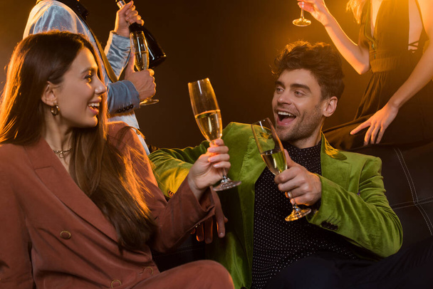happy couple holding glasses of champagne while sitting on sofa near friends on black - 写真・画像
