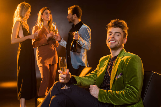 man sitting on sofa and holding glass of champagne near multicultural friends on blurred black background - Foto, immagini