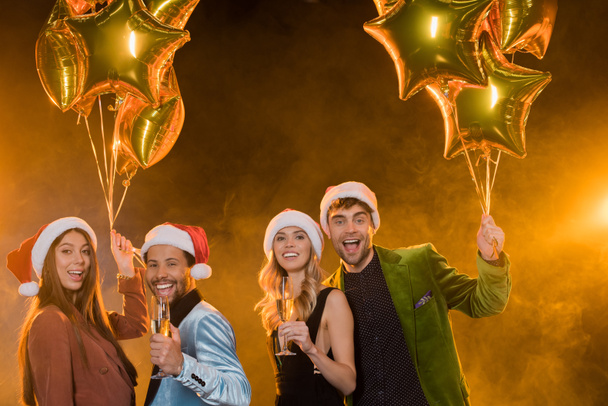 uśmiechnięci wielokulturowi przyjaciele w kapeluszach Mikołaja trzymający kieliszki z szampanem w pobliżu balonów na czarno  - Zdjęcie, obraz