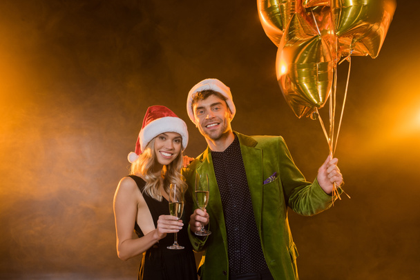smiling couple in santa hats holding glasses with champagne and balloons on black - Foto, afbeelding