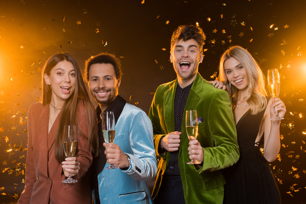 smiling interracial friends holding glasses with champagne near falling confetti on black - Foto, Bild