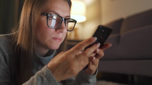 Nő szemüveges fekszik a padlón, és használja okostelefon este - Felvétel, videó