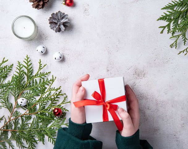 Dziewczyny trzymające biały prezent świąteczny z czerwoną wstążką na jasnoszarym stole. Boże Narodzenie tło z drzewa thuja, świece stożki i zabawki Boże Narodzenie. Widok z góry i przestrzeń kopiowania - Zdjęcie, obraz