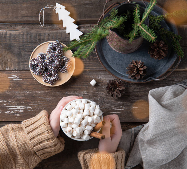 Hands keep christmas mug of cacao and marshmallow with cinnamon on an old brown wooden table with bokeh, cookies, cones and fir. New year mood. Top view - Photo, Image
