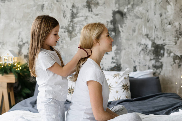 Profile view of little daughter braids hair and making hairdo for her smiling mother. Mom and kid girl playing together, sitting on bed and spending morning at home - Foto, Bild