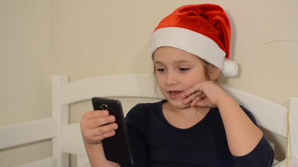 Little adorable girl child in christmas red santa claus hat talking by video call on smartphone - Metraje, vídeo