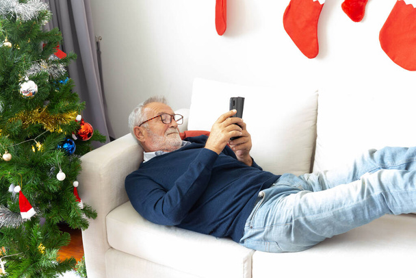 Старший пенсионер расслабиться играть свой мобильный телефон на диване после украшения елки - Фото, изображение