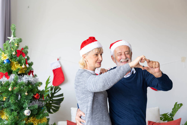 Couple de retraités heureux de danser et de faire un cœur avec leurs mains dans la vie qui décoré par arbre de Noël pendant la période de Noël - Photo, image