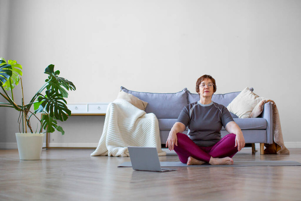 Photo of a senior woman meditaing in front of laptop monitor. Online yoga lesson. - Photo, Image