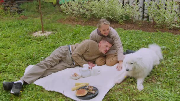 Een jong stel een man en een vrouw op een picknick in de tuin met hun witte hond in slow motion - Video