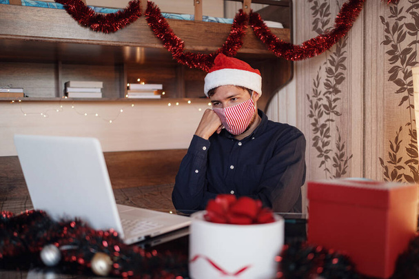 Maskeli mutsuz bir adamın portresi noel süslerinin yanında oturuyor ve hediyeler sipariş ediyor. Yeni yıl alışverişi. Mutlu Noeller Covid 19 Coronavirüs Sosyal Mesafe Konsepti. - Fotoğraf, Görsel