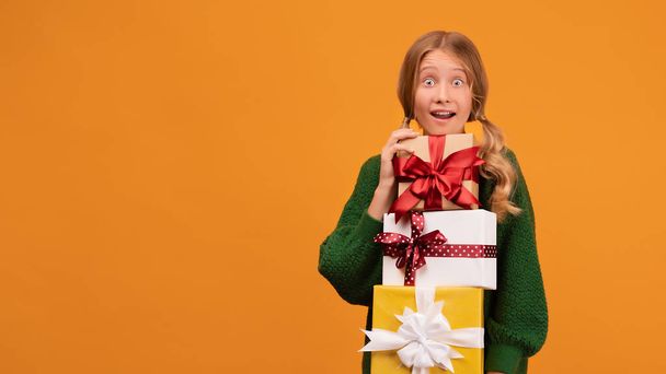 Изображение очаровательной блондинки 12-14 лет в теплом зеленом свитере с большим количеством подарочных коробок. Студийный снимок, жёлтый фон, изолирован. Новогодний женский день рождения - Фото, изображение
