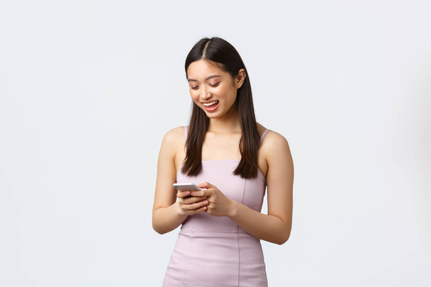 Luxusní ženy, párty a dovolená koncept. Mladý atraktivní asijské dívka ve večerních šatech textové přítel, usmívat se na obrazovce mobilního telefonu, zprávy nebo pomocí aplikace sociálních médií - Fotografie, Obrázek