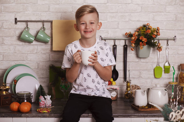 Un beau garçon blond est assis sur la table de la cuisine avec une grande tasse blanche à la main. Photo drôle - Photo, image