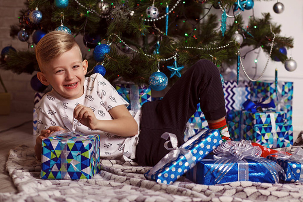 Mignon petit garçon est assis avec un cadeau près du sapin de Noël. L'enfant est heureux avec le cadeau du Nouvel An. Bonne année. Portrait. Photo drôle - Photo, image