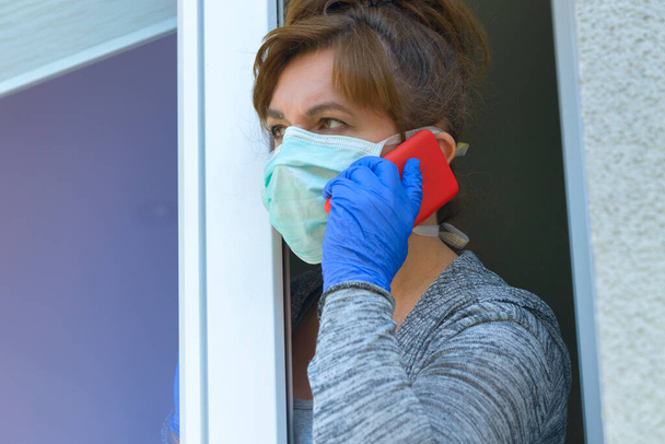 Nainen, jolla on suojaava kasvonaamio ja joka katsoo ulos kotinsa ikkunasta koronaviruksen covid-19-epidemian vuoksi. Kotikaranteeni ja eristäytyminen - Valokuva, kuva