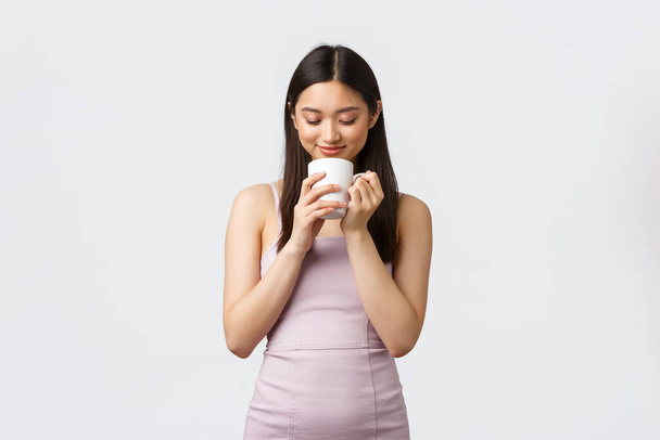 Lifestyle, people emotions and glamour concept. Sensual and romantic smiling asian woman, looking inside coffee cup dreamy, thinking, having nice morning moment, white background - Fotoğraf, Görsel