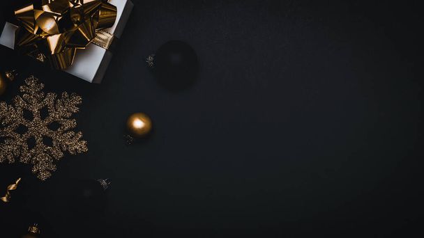 Kerstcadeau. Wit cadeau met gouden strik, gouden ballen en sprankelende lichtslinger in kerstversiering op donkere achtergrond voor wenskaart. Kerst achtergrond met ruimte voor tekst - Foto, afbeelding