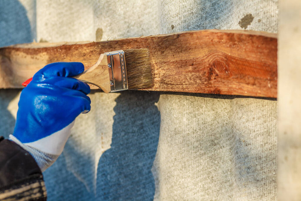 Une main d'homme avec un gant bleu tenant un pinceau appliquant de la laque de bois. Le menuisier teint une planche de bois avec un pinceau à la main avec une peinture mordante. - Photo, image