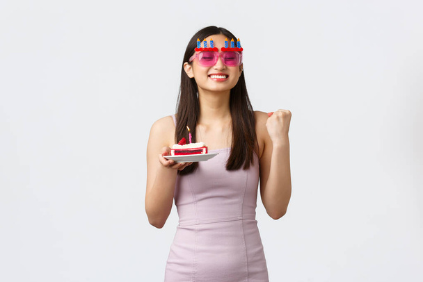 Концепція святкування, вечірки та свята. Весела мила сподіваюся азіатська дівчина в смішних окулярах і одязі, вкоренившись, заохочує себе до позитивного настрою, тримає шматочок торта і робить побажання на день народження
 - Фото, зображення
