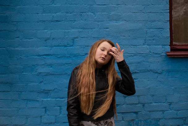 Portret uroczej brunetki nastolatki w skórzanej kurtce na tle niebieskiej cegły ściany. Przytulność, moda, styl. - Zdjęcie, obraz