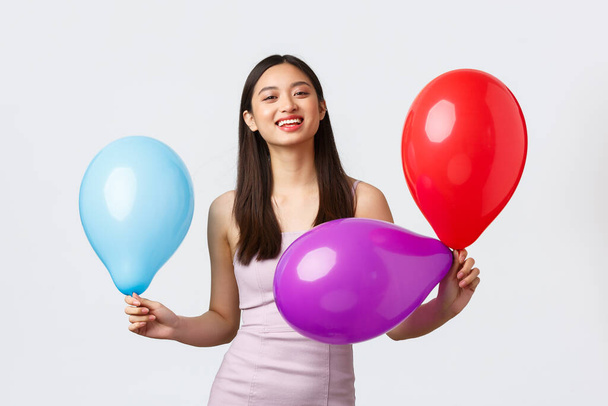 Feste, Feiern und Feiertage Konzept. Fröhlich lächelnde asiatische Frau im Abendkleid, fröhlich lachend, Geburtstag feiernd, bunte Luftballons in der Hand, weißer Hintergrund - Foto, Bild