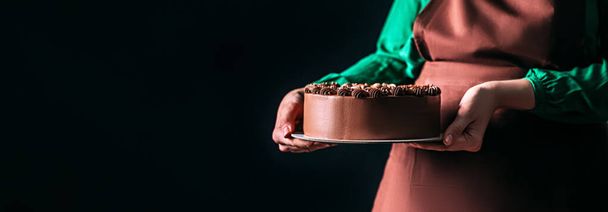 Mujer panadera en delantal sosteniendo un pastel de chocolate sobre un fondo oscuro. Uniforme de trabajadores en la cocina. Lugar para el texto. - Foto, imagen