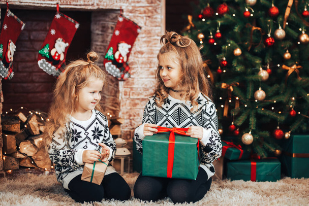 Két nővér egy dobozt tart egy ajándékkal egy zöld csomagban. Karácsonyi hangulat. A háttérben egy díszített karácsonyfa és egy kandalló karácsonyi zoknival. - Fotó, kép