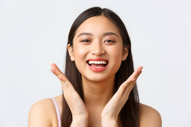 Concepto de publicidad de productos de belleza, moda y maquillaje. Primer plano de hermosa chica asiática mostrando sus productos para el cuidado de la piel en la cara, encantado con el maquillaje, sonriendo optimista - Foto, imagen