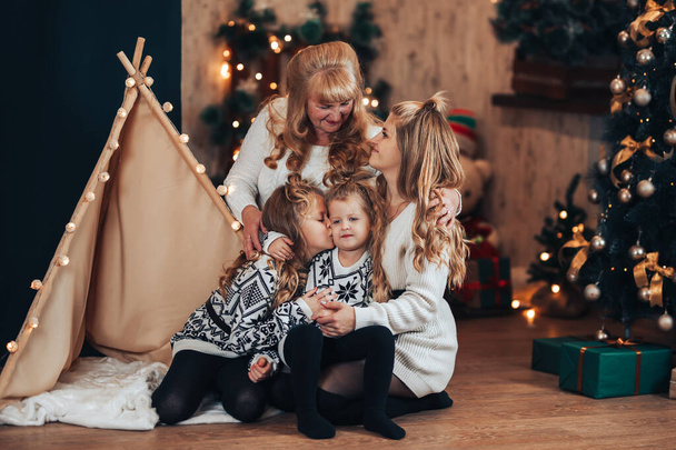 母親と祖母を持つ子供たちは、テントやクリスマスツリーに抱きついて座っています。クリスマス気分。家族全員で休日を祝う. - 写真・画像