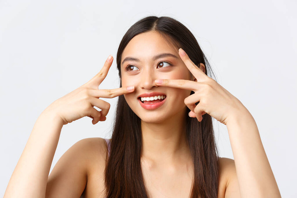 Concetto di bellezza, moda e trucco pubblicità prodotti. Close-up ritratto di bella sognante ragazza asiatica guardando a sinistra felice, mostrando segni di pace e kawaii sorridente, sfondo bianco - Foto, immagini