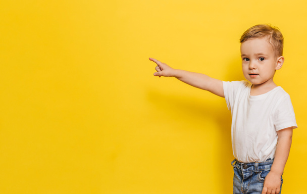 A little boy on a yellow background points to the background. Copy space. - 写真・画像
