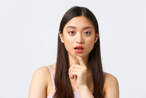 Schoonheid, mode en mensen emotie concept. Close-up portret van geïntrigeerd aziatische vrouw luisteren, aanraken kin en open mond terwijl op zoek nieuwsgierige camera, witte achtergrond - Foto, afbeelding
