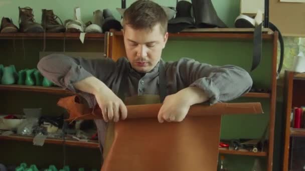 Egy cipész tanonc egy darab valódi bőrt tekerget egy tekercsbe. A háttérben, állványok üres cipők. - Felvétel, videó