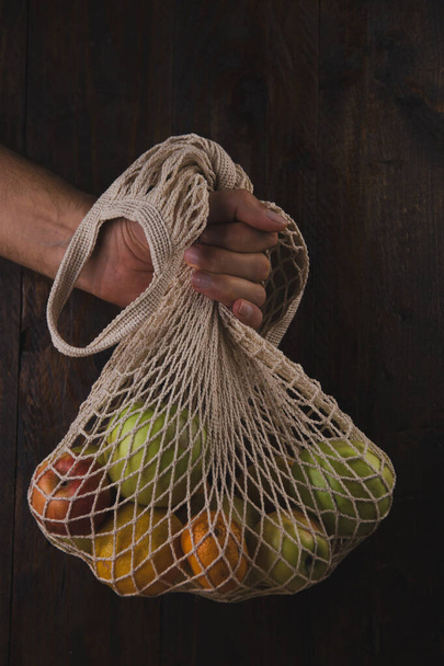 Mano de hombre con bolsa de red con frutas orgánicas contra una tabla de madera oscura. Ecológico, cero residuos, concepto libre de plástico - Foto, imagen