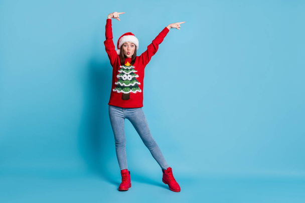 Full body size photo of cute adorable lady raise hands direct indicate empty space lips wear santa x-mas headwear red ornamented pullover jeans boots isolated blue color background - Fotó, kép
