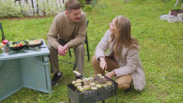 A woman on the grill in the country roasts zucchini, a man helps her, they talk and laugh - Footage, Video